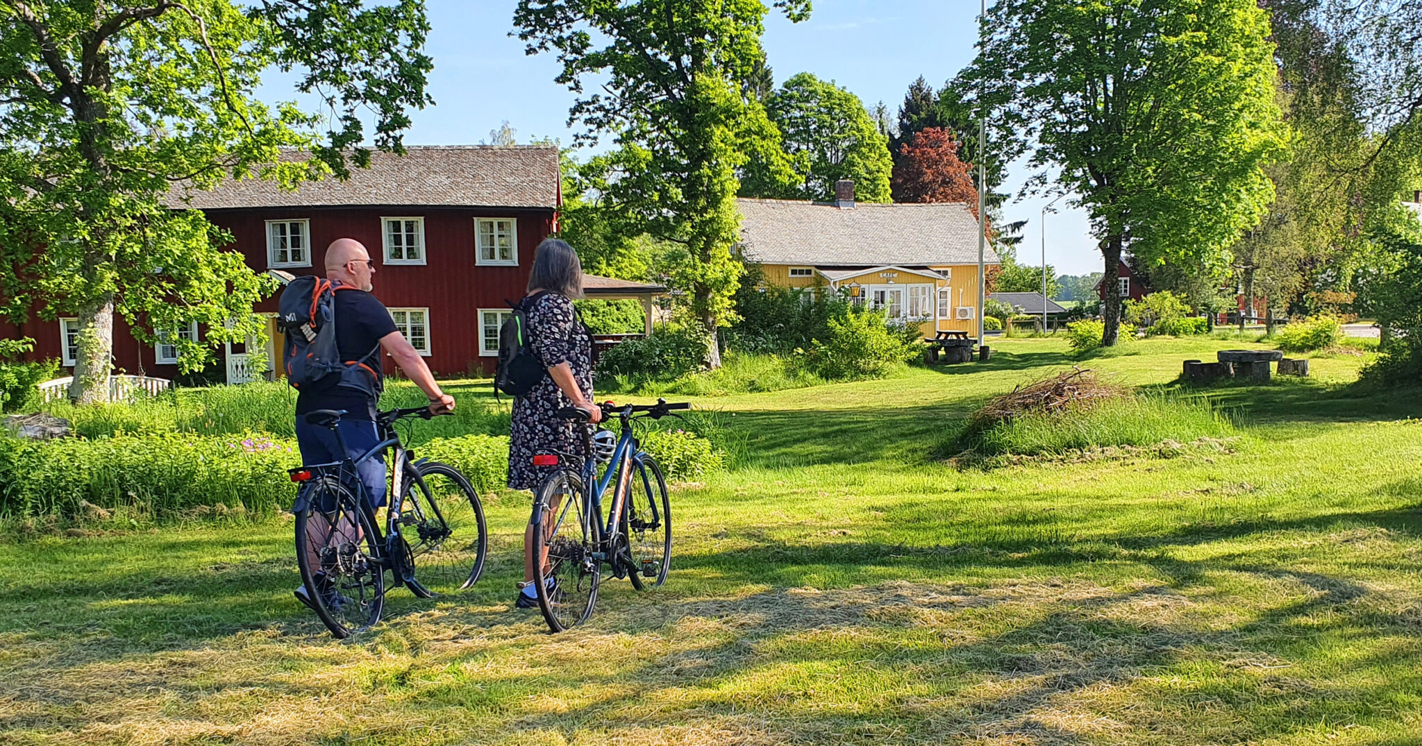 Två personer med cyklar och ryggsäck går in i ett lummigt område med små hus.
