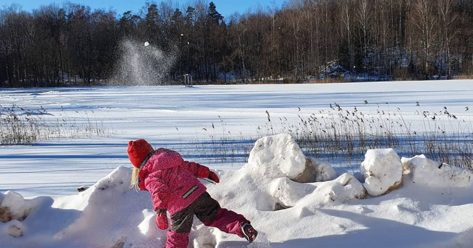 Boy in pink winter overalls throws snow at a ice covered lake.